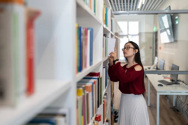 コンピュータを備えた読書室 図書館の本棚から本を取る女性学生を勉強しながら 科学文学を探している眼鏡の若いアジアの大学生の女の子 文学検索 — ストック写真