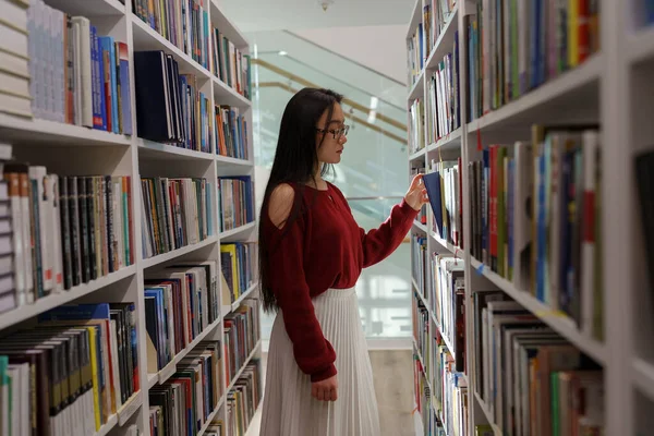 Thoughtful asian student girl searching materials for educational research in college library. Young chinese woman choosing book for reading in bookstore, selective focus. Literature and education