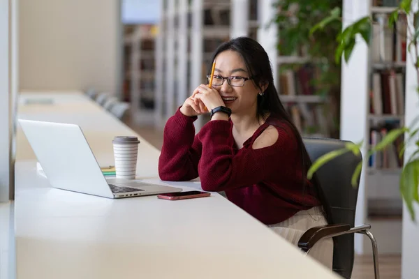 Happy asian girl studying in library. Young chinese woman in glasses with laptop and notebook sit at desk in modern coworking space cheerful enjoy online education, freelance work or remote occupation