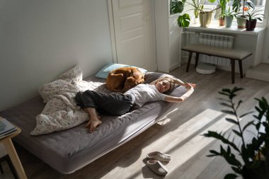 Lazy weekend. Relaxed Scandinavian 40s woman pet owner lying in bed with dog in early morning, relaxing after waking up, happy middle-aged single female wearing pajama stretching body after sleeping