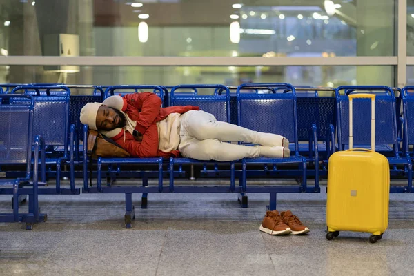 Young African American man is napping while waiting for flight, feel cold. Black guy lying on chairs in airport lounge, can not sleep on an uncomfortable bench, freezing. Long night connection.