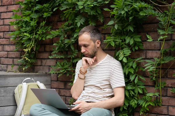 Thoughtful caucasian millennial man wear casual outfit distance working on laptop. Focused young hipster male reading news on computer. Unshaven guy green calm workplace sitting