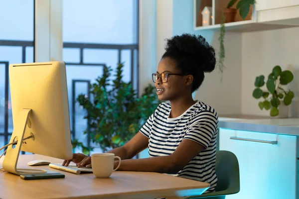 Happy young african millennial woman wearing eyeglasses working remotely at cozy at home office, joyful black student girl looking at computer screen watching live webinar, enjoying remote learning