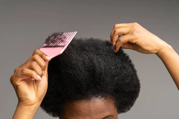 Close up of African American woman using pink plastic comb over studio gray wall background. Unrecognizable black girl brushing curly trick hair, Haircare, daily routine concept.