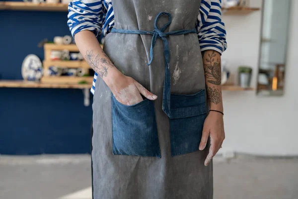 Female ceramist put hand at pocket after working with raw clay in pottery studio. Young student girl or master posing art dirty apron at her studio. Pottery business and workshop concept