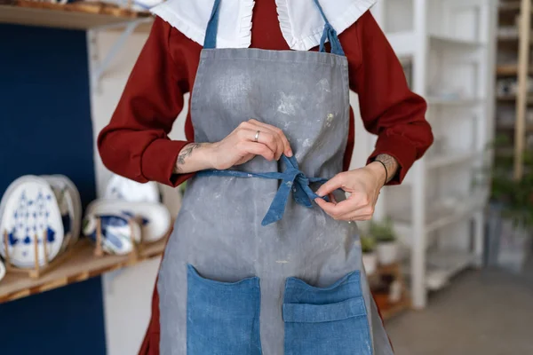 Female ceramist tying her apron before working with raw clay in pottery studio. Young student girl or master posing art dirty apron at her studio. Pottery business and workshop concept