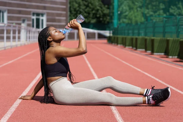 Attractive fit young female in sport wear rest on street sports training area. Black fitness girl replenishes energy after running under scorching sun. Athletic human resting from hard sports activity