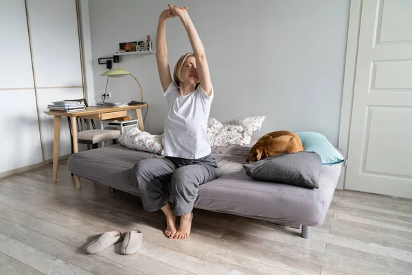 Good morning. Calm peaceful middle-aged woman living alone with dog sitting with closed eyes and raised arms on sofa in bedroom, waking up alone, stretching arms, female in sleepwear getting up early