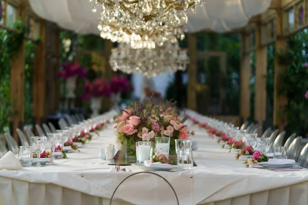 Wedding banquet table with pink rose flowers decoration, crystal chandelier in summer garden, selective soft focus, blurred background. Table set for holiday, event, party in outdoor restaurant.