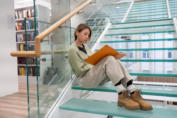 Curious middle-aged woman looks into book with orange cover sitting on modern glass stairs step. Focused female student in casual outfit enjoys reading interesting story in large university library