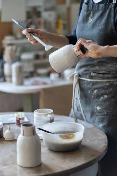 Crop photo of woman artisan making earthenware vase. Female skilled worker hands prepare clay item with brush for painting in big workroom. Young craftswoman enjoys creating things from clay closeup