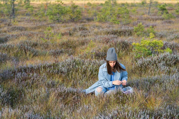 Young woman relaxes sitting on autumn swamp grass. Stylish girl in hipster hat collects ripe cranberries into small container. Female travels outside city for recreation and unity with nature