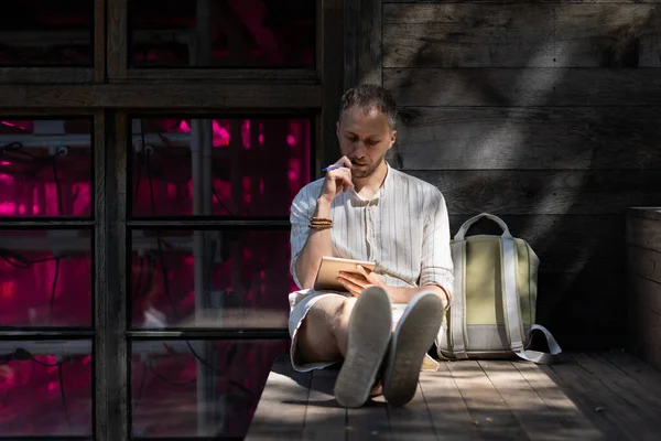 Young man student wearing white shirt sits on wooden stairs step by bag outdoors. Stylish guy makes notes in paper notebook preparing for examination sitting against building wall in rays of sunlight