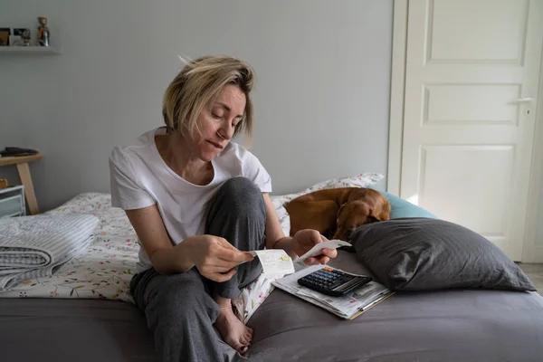 Unhappy middle-aged woman wants to cry after looking at debts of apartment utility bills. Lonely mature woman in nightwear loses motivation and life sense sitting on bed near sleeping dog closeup