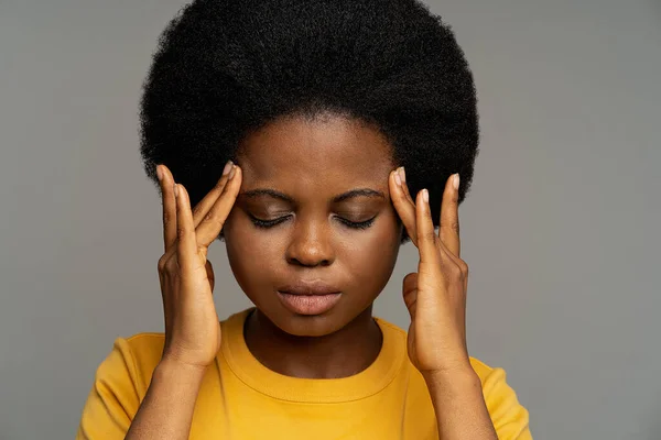 Tired black young woman in yellow t-shirt feels lack of concentration after long work in office. African American lady with kinky hair closes eyes putting fingers on temples on grey background closeup