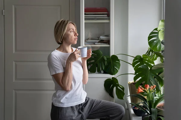 Tranquil Middle Aged Female Enjoys Hot Drink Mug Standing Windowsill — Zdjęcie stockowe