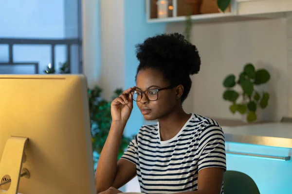 Focused black student girl wearing eyeglasses looking at computer screen, concentrated young African woman watching educational webinar, reading article online at home. Distance learning and education