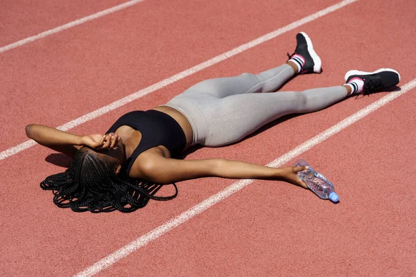 Black female beginner feels powerless after running long distance on hot summer day. Exhausted African American woman lies on red track holding bottle of water and covering eyes with hand from sun