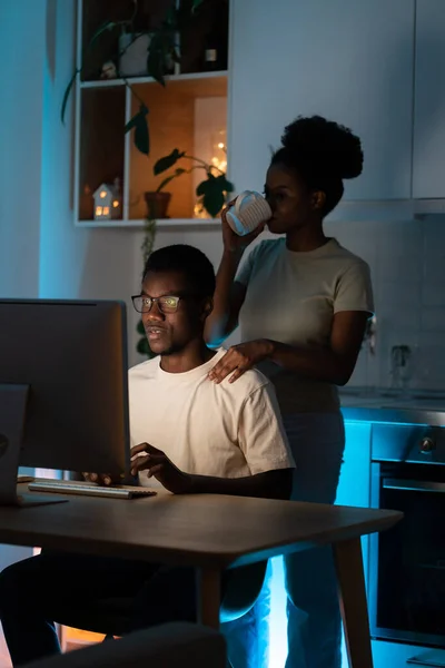 Young African American couple stays up late due to work on new profitable project. Woman drinks coffee putting hand on shoulder of focused man in glasses sitting at kitchen table near computer closeup