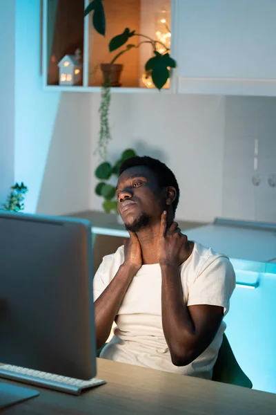 Black freelancer in white t-shirt feels tired of staying up late at work and developing new project. African American man kneads neck with hands sitting at wooden table by computer in kitchen closeup