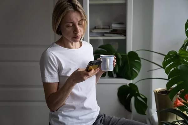 Middle Aged Blonde Woman Scrolls Social Nets Smartphone Holding Mug — Stockfoto