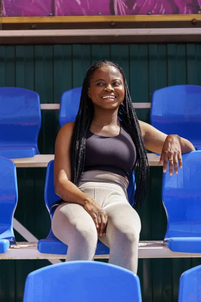 Cheerful African American woman takes break from workout sitting on blue seat on city stadium and smiling. Young sportswoman with long dark braids enjoys watching training of local football team