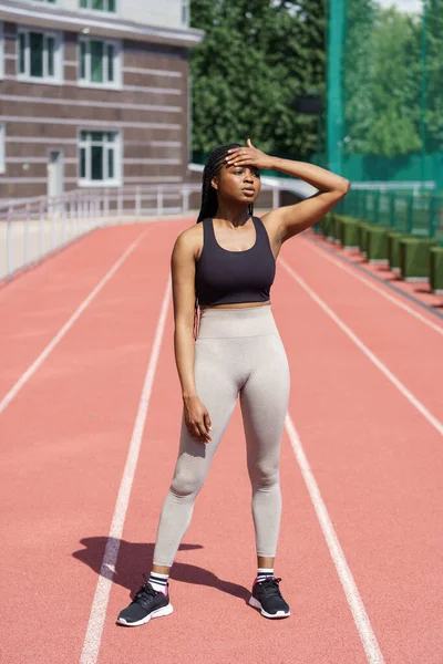 Tired black lady in sportswear puts hand on forehead standing on red track of big city stadium. Young fit African American woman stops to take break after running long distance on sunny summer day
