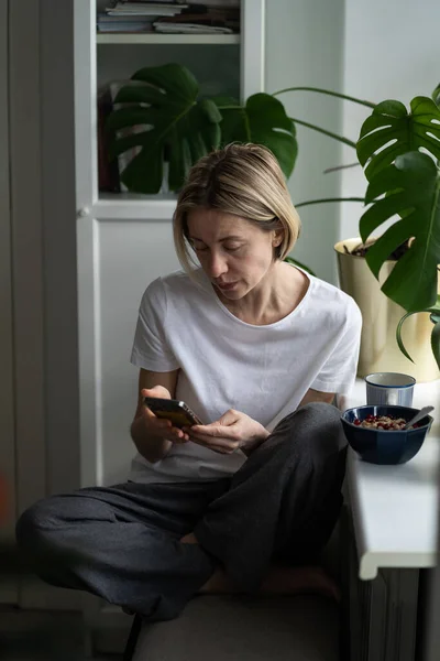 熟女はスマートフォンを使ってインターネットで空きを検索している窓の近くに座っています 希望に満ちた中年ブロンド女性座っていますによってWindowsillとともにフードウォッチング仕事提案上の特別なウェブサイトクローズアップ — ストック写真