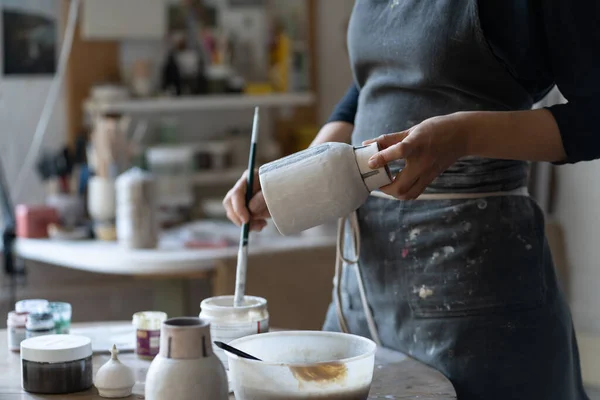 Crop photo of woman artisan making earthenware vase. Female skilled worker hands prepare clay item with brush for painting in big workroom. Young craftswoman enjoys creating things from clay closeup