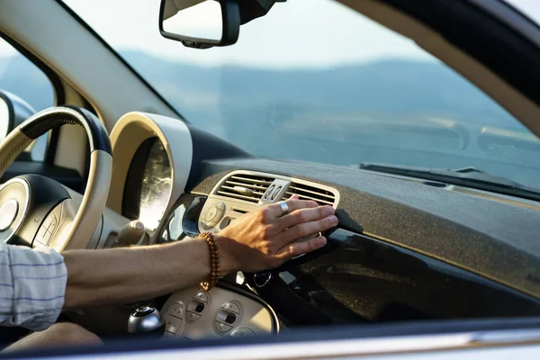 Driver goes on road trip sitting at steering wheel and driving in rural area on hot summer day. Man puts hand on modern panel vent to check cold air flow and operation of ventilation system closeup