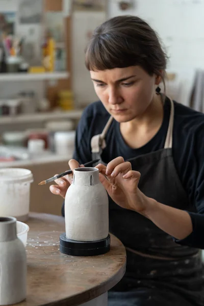 身穿黑色围裙的年轻女工匠与陶器一起工作 她们坐在明亮窗户旁边的车间的圆桌旁 布鲁内特女人喜欢在陶器工艺工作室里画手工制作的陶瓷花瓶 — 图库照片