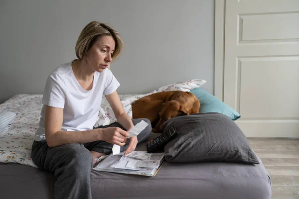 Depressed middle-aged woman wearing old nightwear thinks about paying utilities looking at bills. Mature lonely female unsatisfied with cost of living sitting on bed with domestic dog closeup