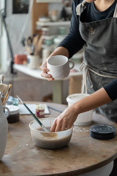 车间里 穿着脏围裙的女人拿着白色陶瓷杯 站在圆木桌旁模糊的背景上 女工匠用手拿着装有油漆的碗 用手工制作的陶器做特写 — 图库照片