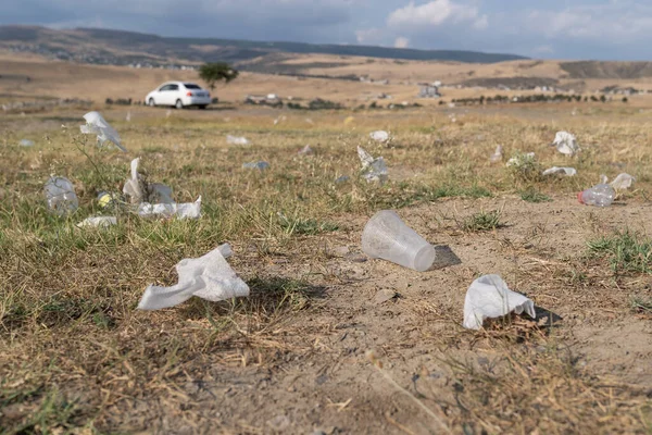 Rubbish from garbage dumb scattered on yellowed grass of meadow causing environmental pollution in wild area. Plastic cups and bags lie in countryside under cloudy sky in summer. Ecological problem