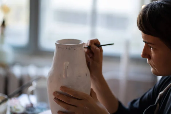 女人们在车间明亮的窗边的桌子边学习手工制作陶器 聚焦的黑发女人在模糊的背景特写下 用刷子在白色陶瓷花瓶上画文字 — 图库照片