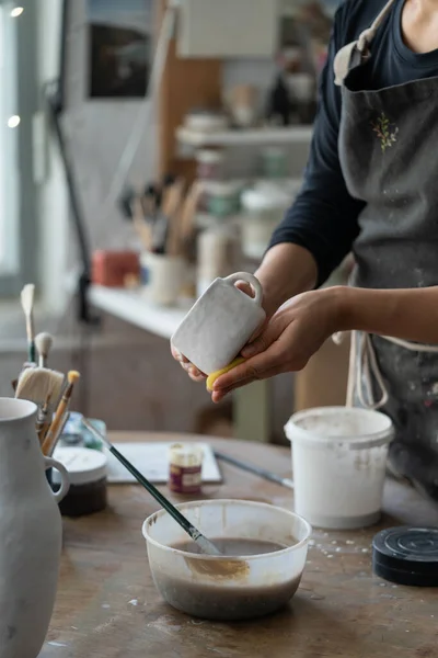 身穿黑色围裙的妇女站在工作室圆木桌旁加工白色瓷杯 女性喜欢在手工制作的陶器中工作 学习烹饪特写专业课程 — 图库照片