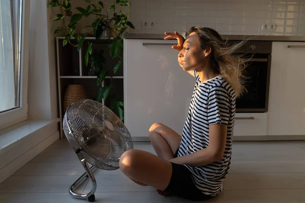 Exhausted young woman refreshing sit near big indoor ventilator blowing cooling fresh air at home on floor bonding. Tired female enjoying conditioner having fun indoors. Summer, heat weather concept