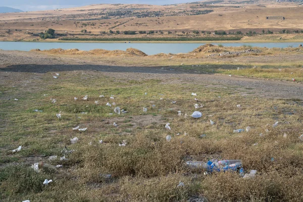 Rubbish from garbage dump spread through meadow in rural area on sunny summer day. Plastic bags and bottles scattered on dry yellowed grass near pond with water in countryside. Environmental pollution