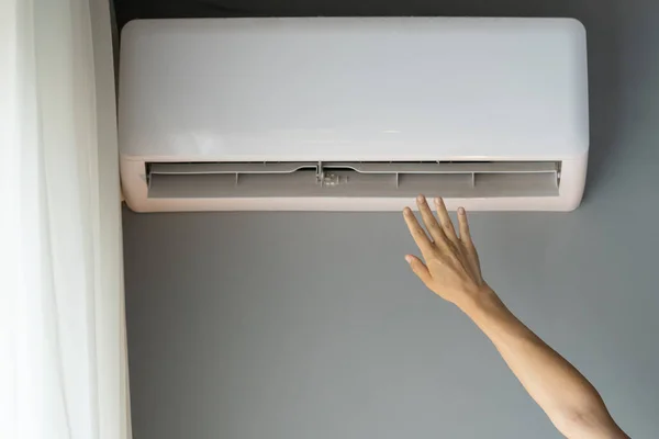 Woman wants to catch stream of warm air stretching hand to device slot at home. Female turns on air conditioner hanging on grey wall to warm up apartment to feel better