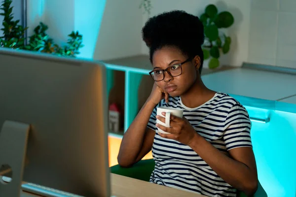 Bored African American entrepreneur thinks about business project drinking beverage from white mug. Black woman wearing glasses looks at computer with thoughtful and serious expression holding mug