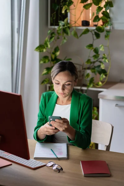 Young business woman in green jacket in home office using smartphone. Cozy workplace next to large window and home plants in pots, remote work. Female worker distracted by social networks