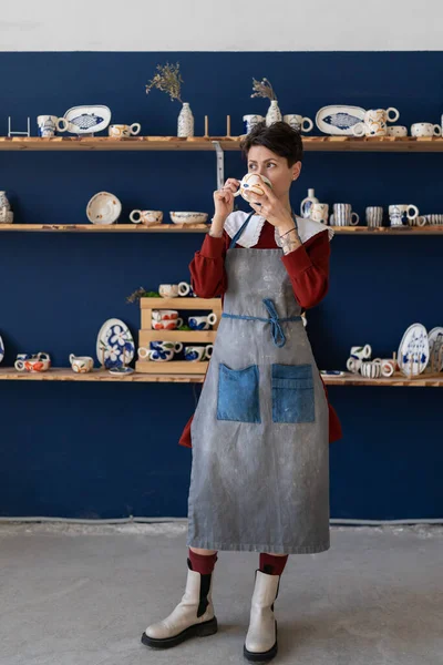 Young creative artisan crafts woman wearing apron drinking morning coffee from handmade clay mug and relaxing while working in her own cozy pottery studio. Small art business concept