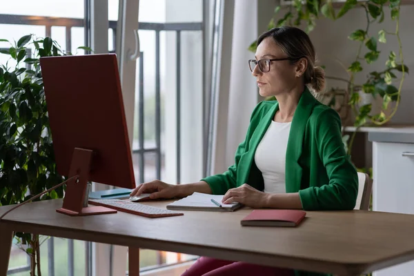 Focused freelancer making future project types information on computer. Young businesswoman wearing glasses learns important details about project and works in cozy home office with green houseplants