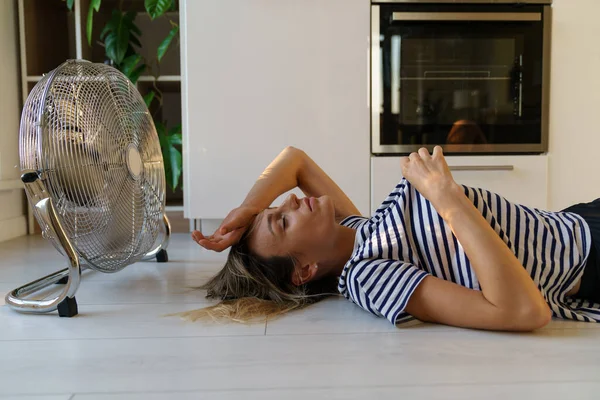 穿着T恤衫的年轻女子躺在电扇旁边的地板上 试图抓住冷空气来降温 躺在地板上汗流浃背的女人讨厌公寓里闷热闷热 — 图库照片