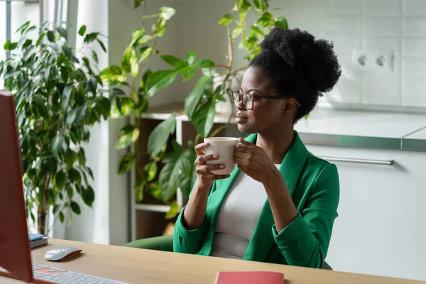 仕事中に休憩を取り ビジネス上の問題を解決する方法を考えながら アフリカ系アメリカ人女性がコーヒーを飲む成功した スタイリッシュなスーツの女の子は自宅のオフィスでコンピュータとテーブルに座って — ストック写真