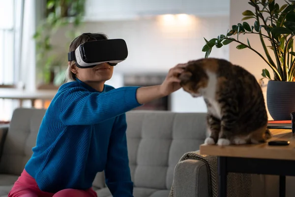 Young woman wearing VR glasses explores world of virtual game at home. Female wearing blue sweater reaches with finger domestic cat sitting on wooden table and strokes calm pet head in real life