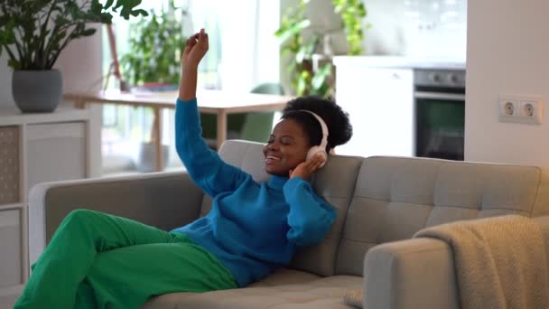 年轻的非洲裔美国女人戴着耳机 坐在客厅的沙发上 享受着最喜爱的作曲和歌曲 — 图库视频影像