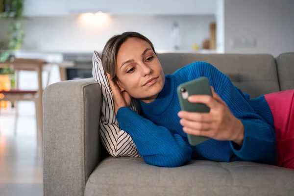 若い女性は ソーシャルメディアのボーイフレンドからの悪いニュースの返信メッセージを読んでスマートフォンを見ています 快適なソファクッションに横たわっている女性は居心地の良いアパートでインターネット滞在について考えています — ストック写真