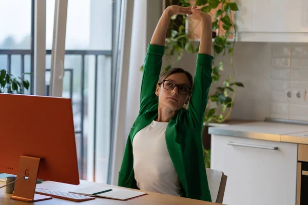 Businesswoman in green blazer and glasses stretches arms after hard work on future project at home workplace. Stylish freelancer takes small break from distant work on computer in modern apartment