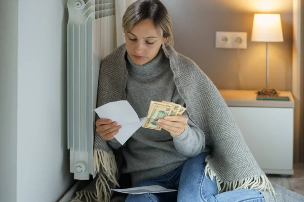 Sad woman leaning on cold central heating radiator looks at bills and holds money. Lady covered with warm blanket sits looking at debts without knowing how to solve problem and prevent bankruptcy